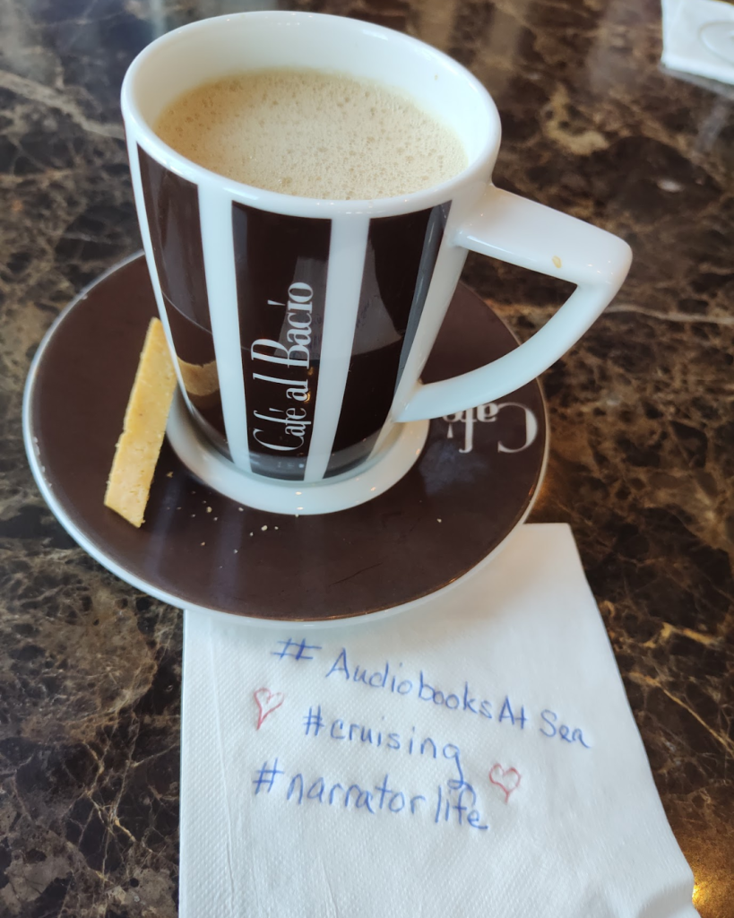 a latte in a brown and white striped coffee cup on a matching saucer, sitting on a napkin that says #audiobooksatsea